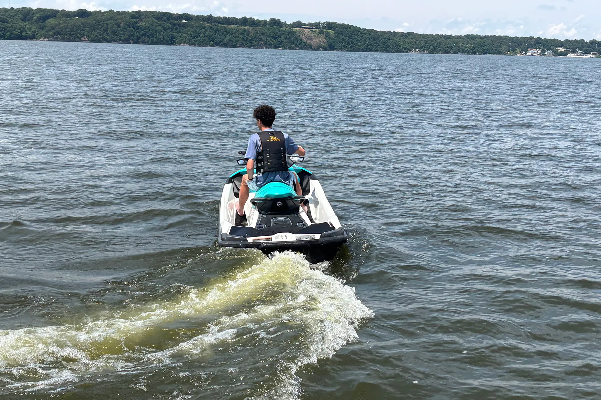 Person riding a teal and white jet ski on a lake with trees in the distance under a cloudy sky.
