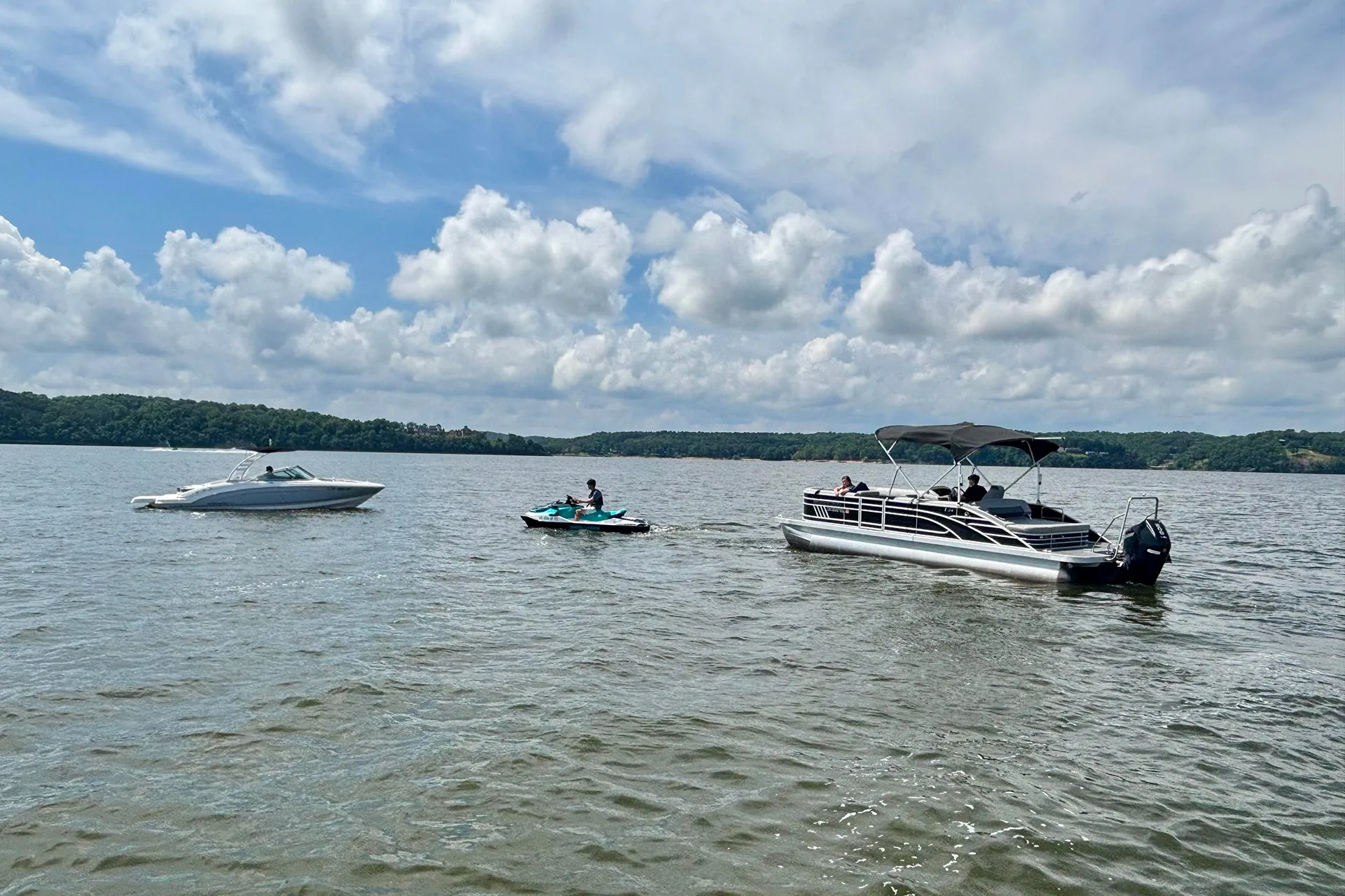 Two boats and a person on a jet ski on a lake under a cloudy blue sky.