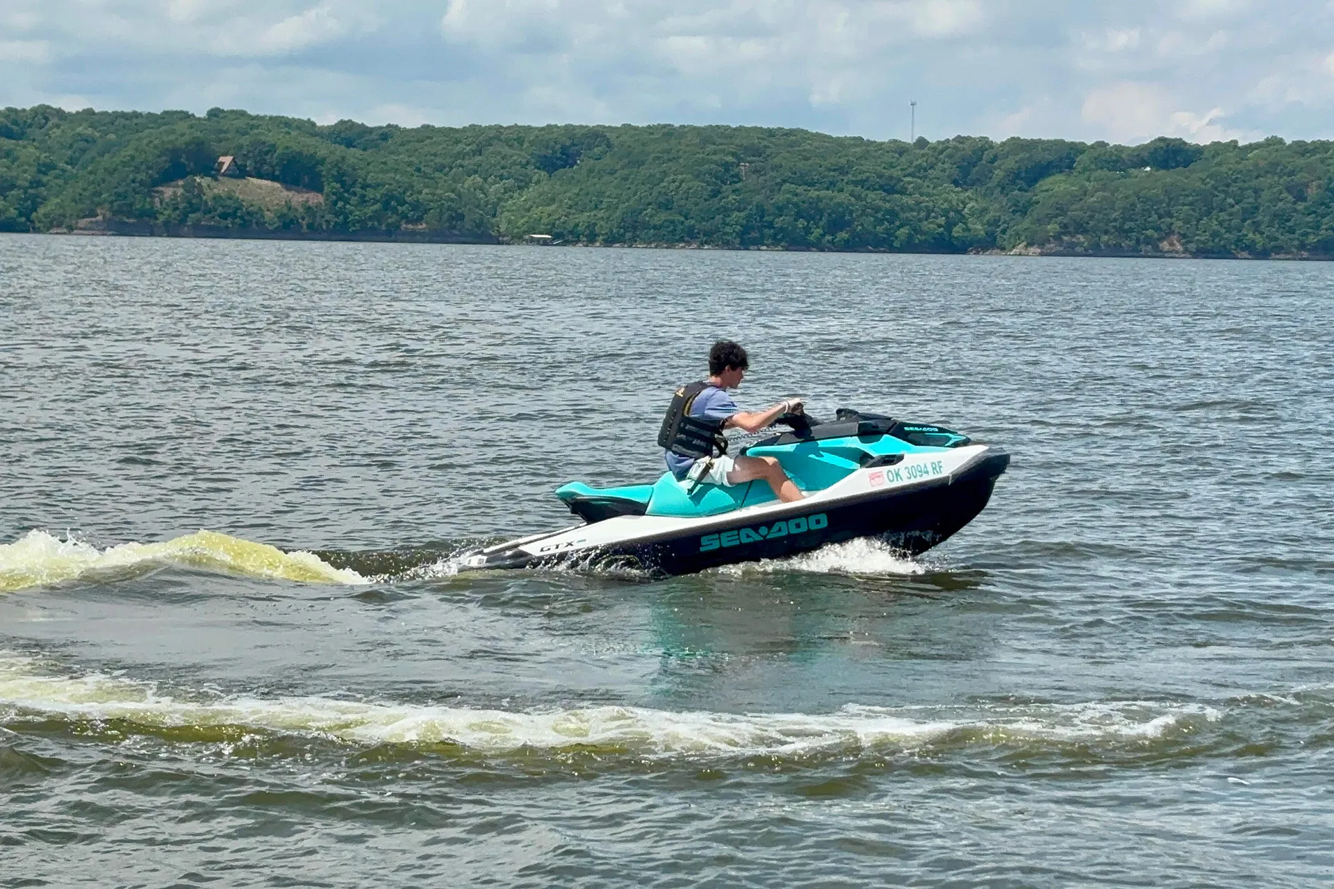 Person riding a teal and white Sea-Doo jet ski on a lake near a forested shore.