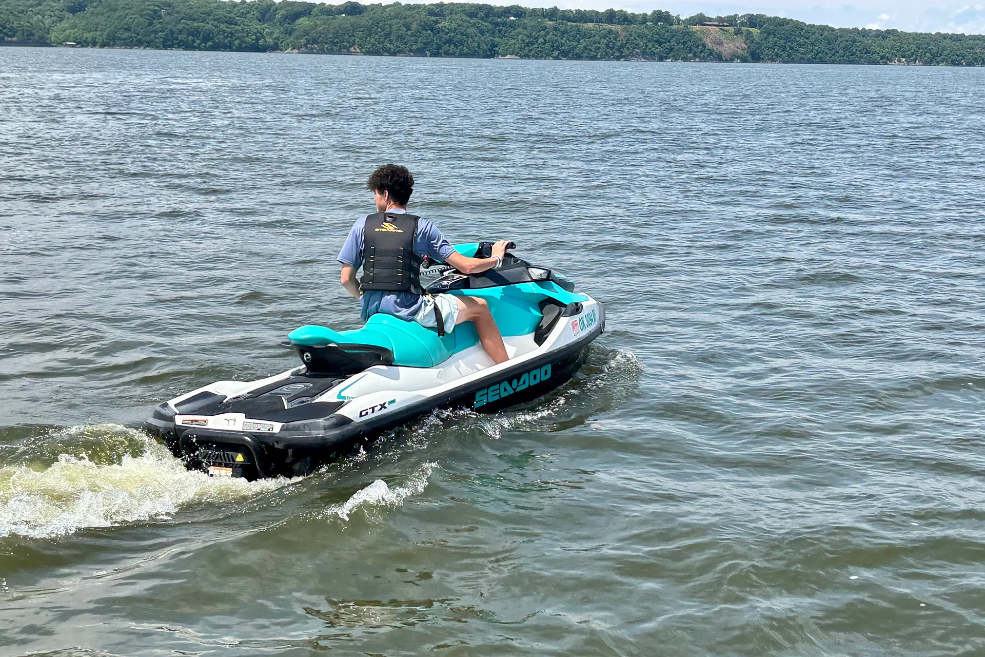 Person riding a teal and white Sea-Doo GTX jet ski on a calm lake with forested shore in the distance.