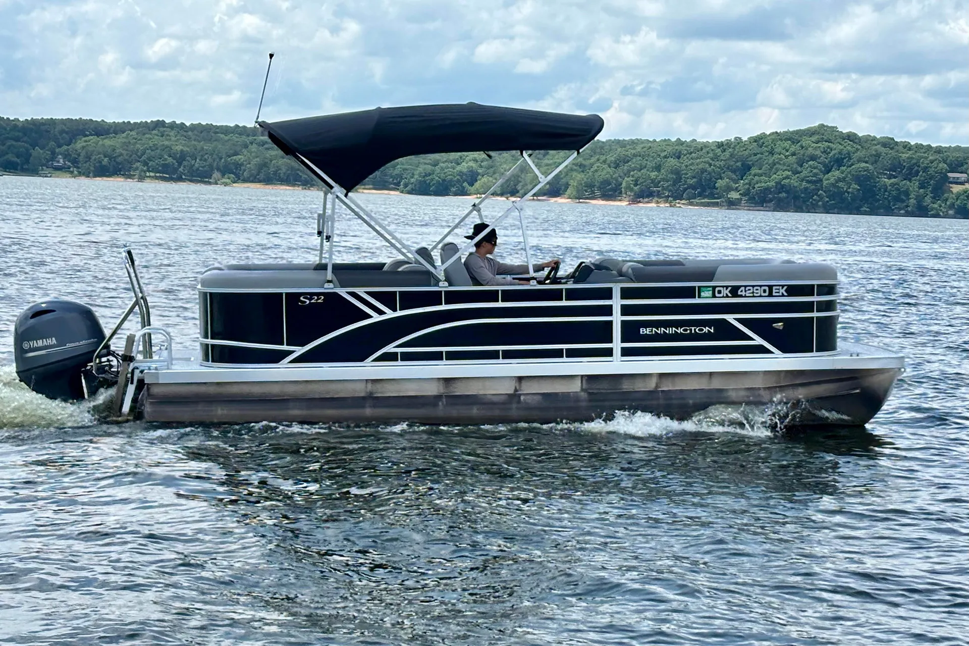 Person piloting a Bennington pontoon boat on calm water with a wooded shoreline in the background.