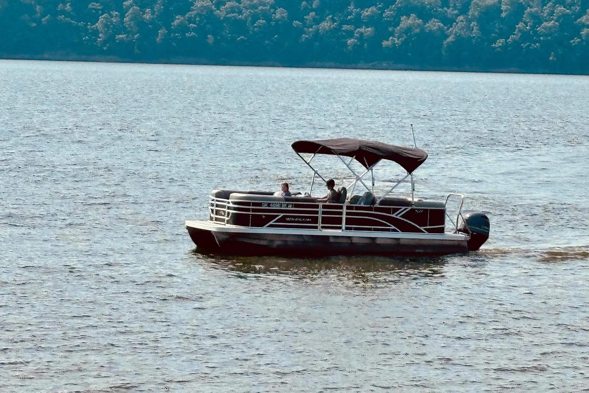 Pontoon boat with two people cruising on a calm lake with tree-lined shore in the background.