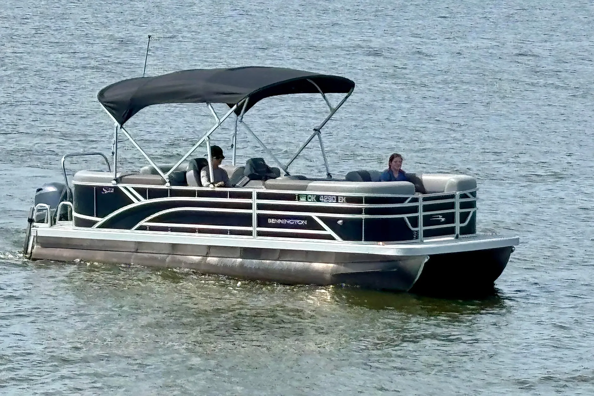 Pontoon boat with black canopy cruising on water carrying two people seated inside.