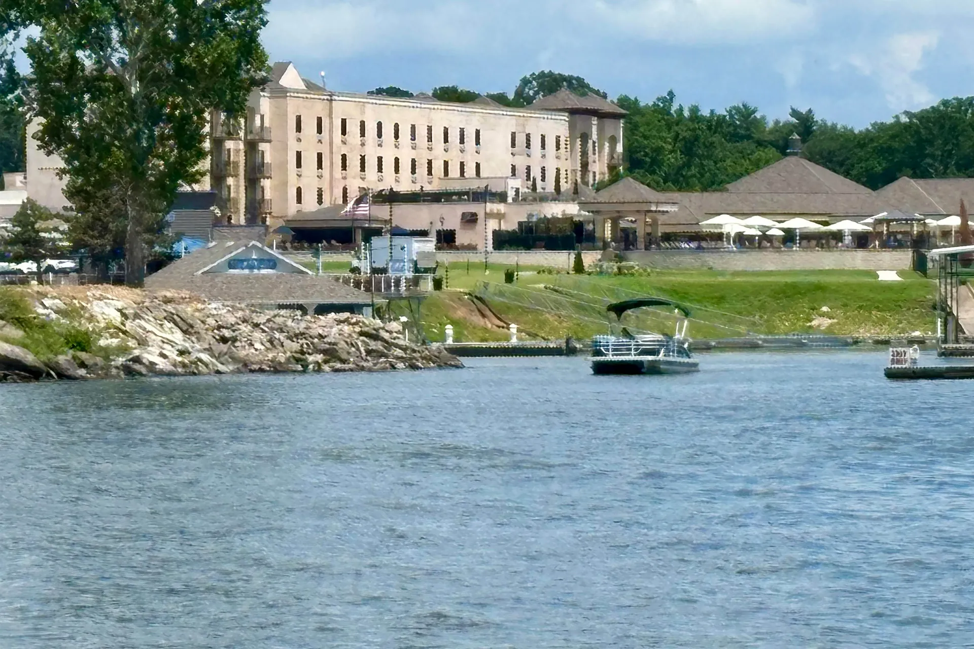 Pontoon boat floating on lake in front of large building, pavilion, and green landscape on shore.