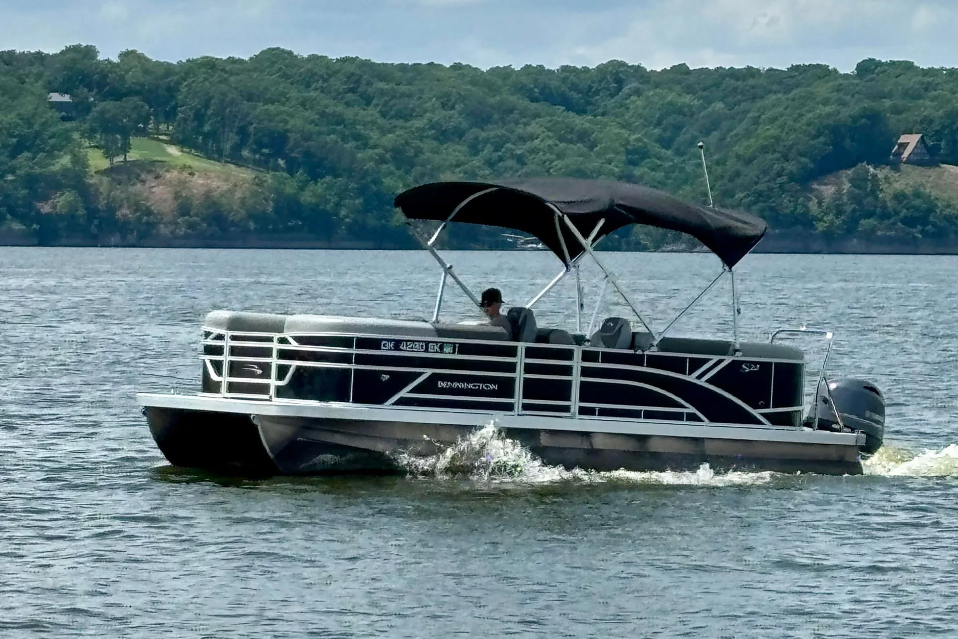 Black Bennington pontoon boat on water with a person seated under a black canopy and green forest in background.