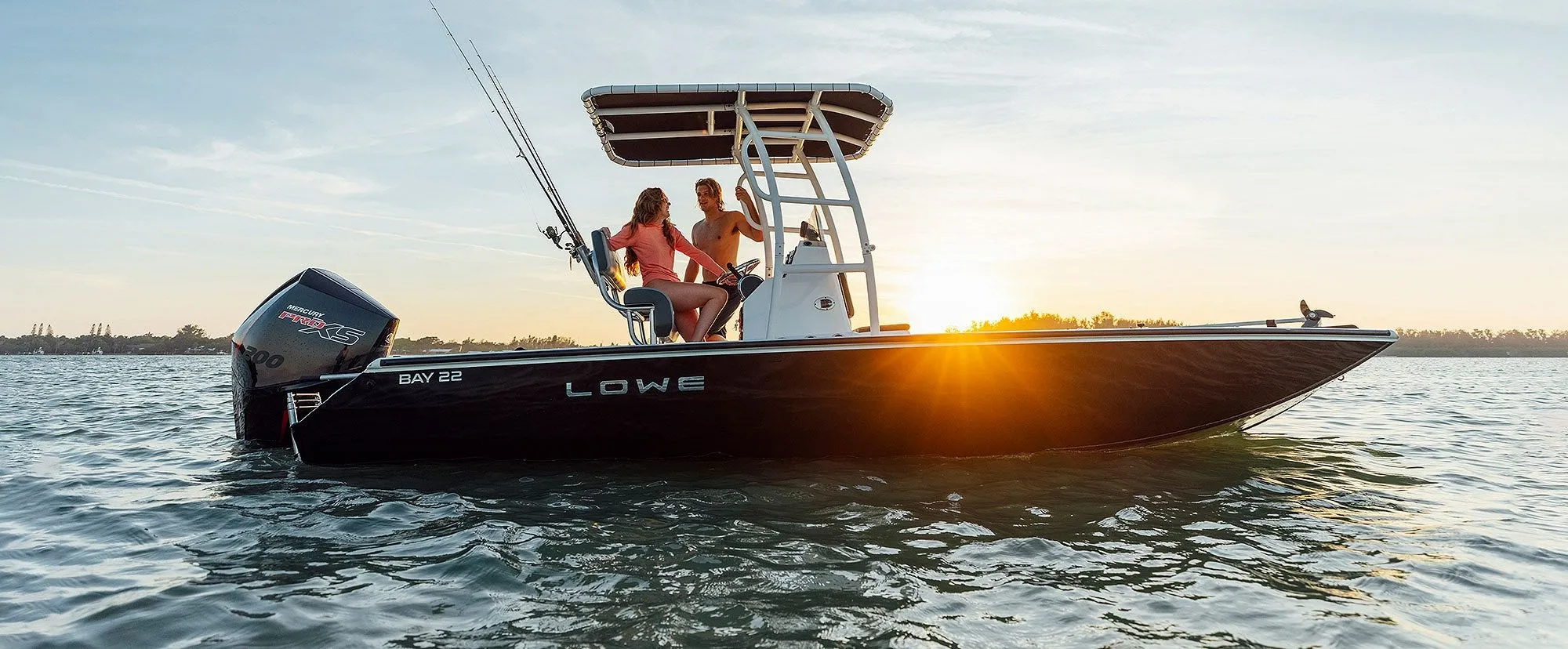 Two people on a Lowe Bay 22 boat with fishing rods during a sunset over calm water.