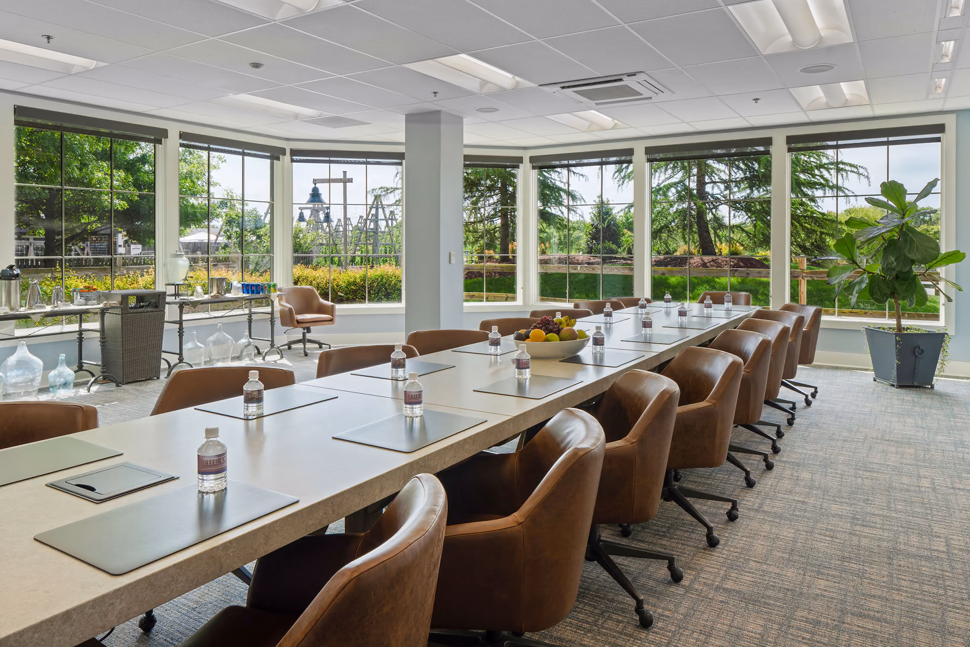 A modern conference room with a long table, brown chairs, bottled water, and large windows overlooking greenery.
