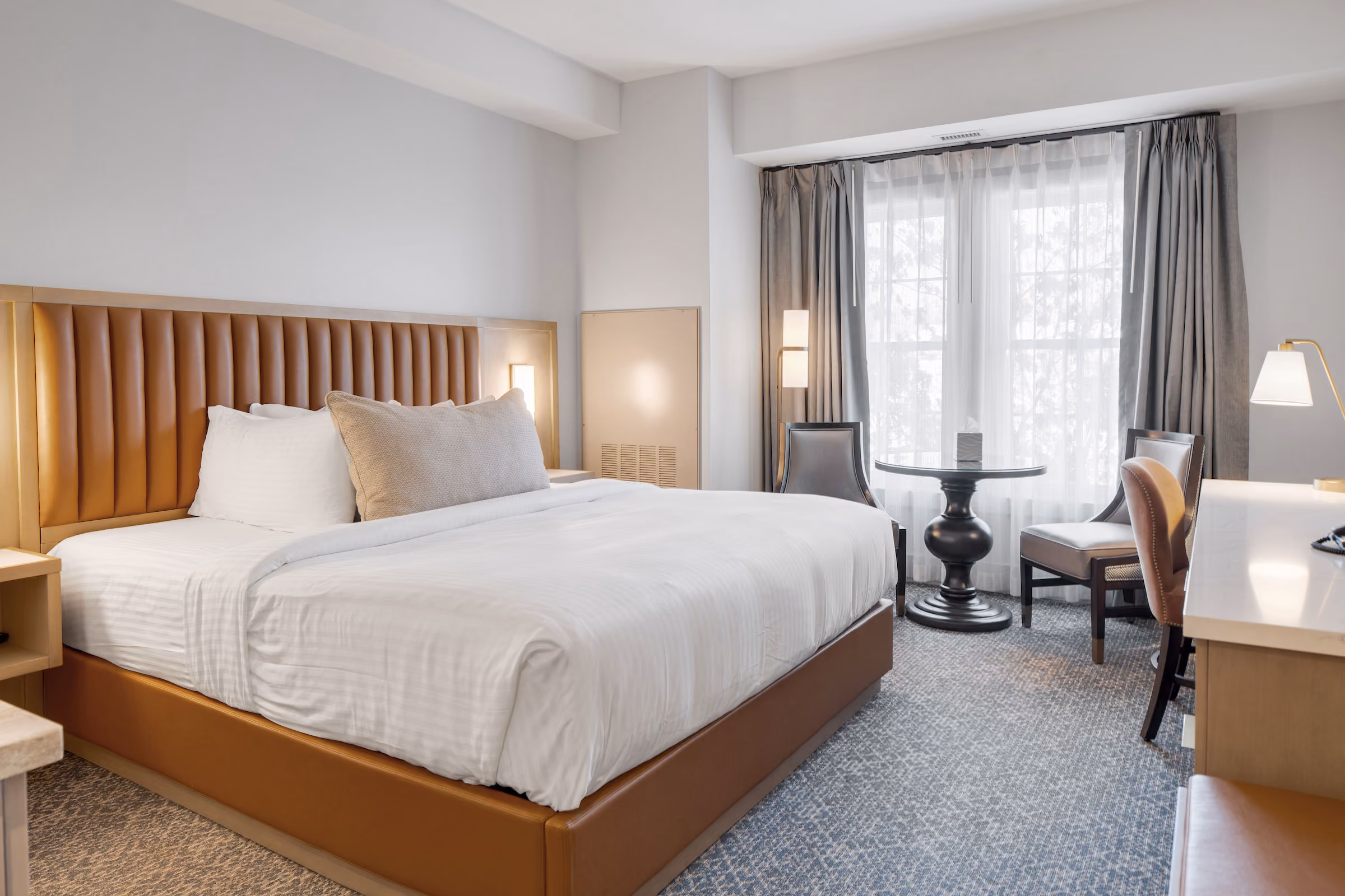 Modern hotel room with a large bed, desk, chairs, round table, and window with sheer curtains letting in natural light.