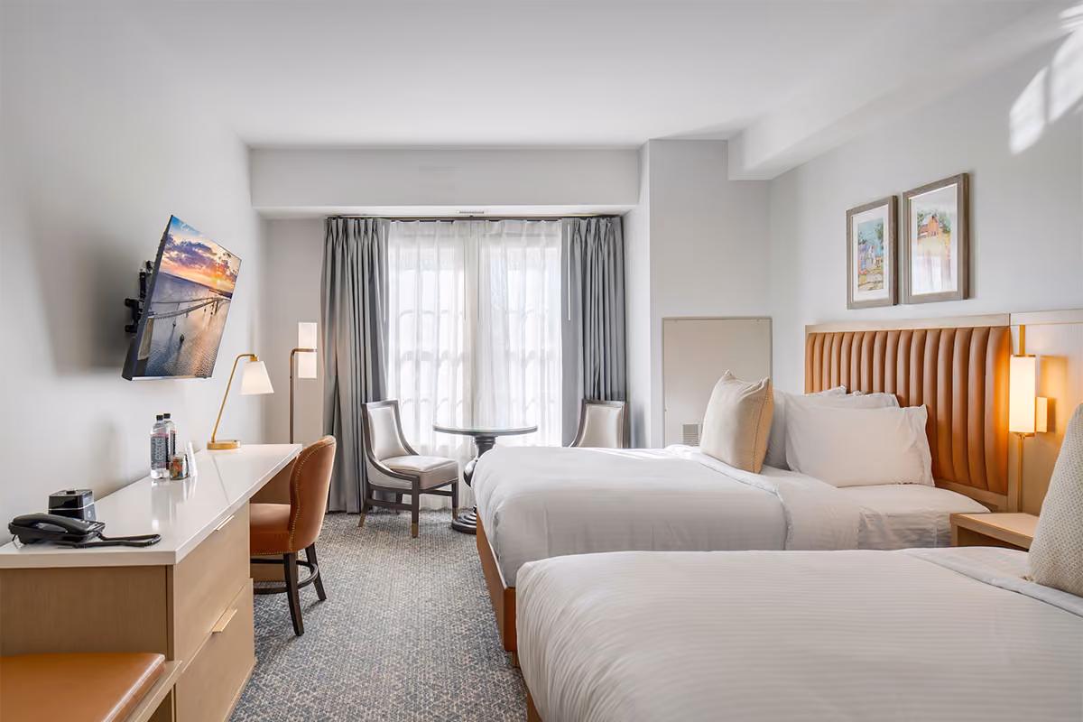 Modern hotel room with two double beds, a desk, flat-screen TV, seating area by window, and neutral decor.