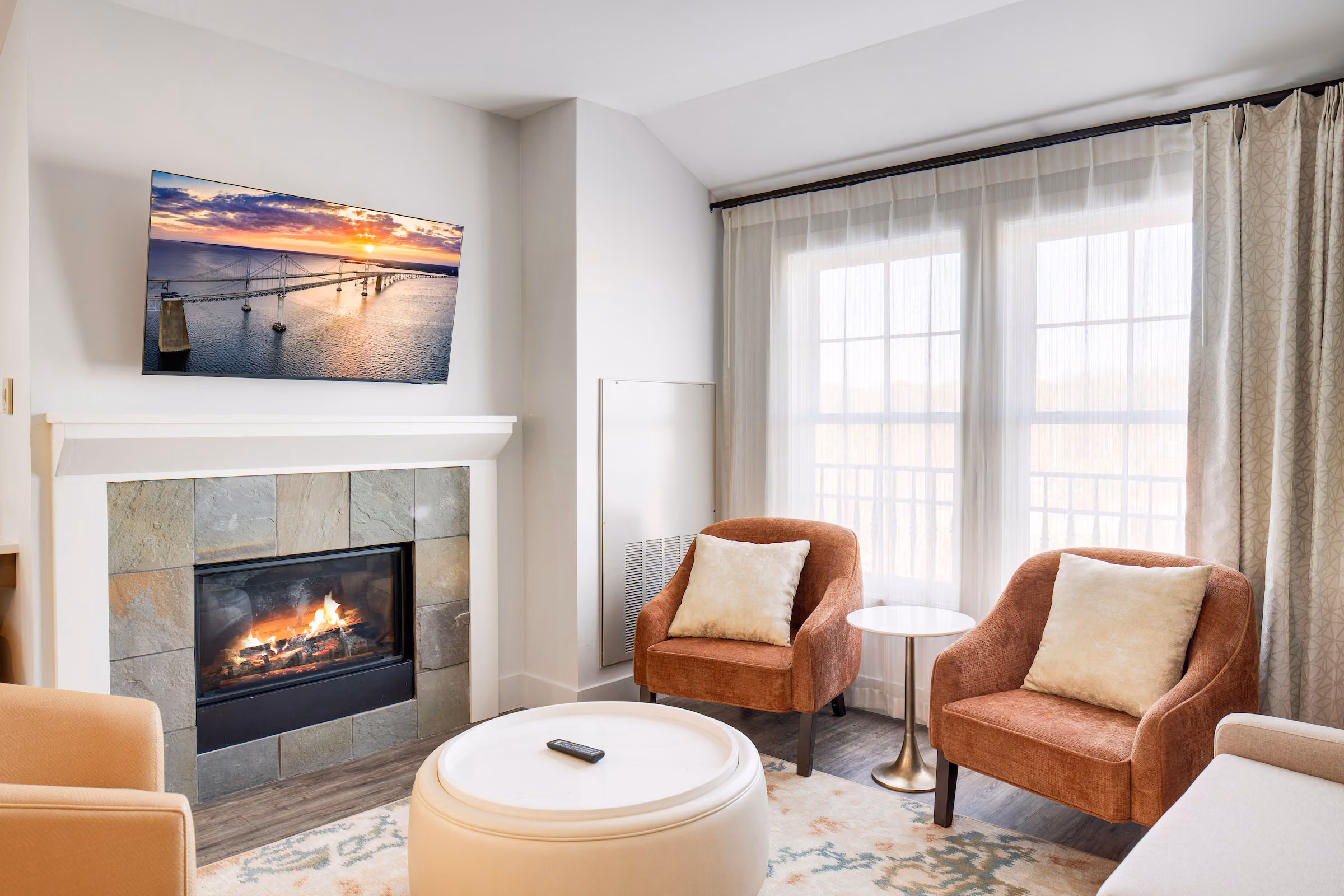 Modern living room with a lit fireplace, two armchairs, a TV above the mantel, large windows, and sheer curtains.