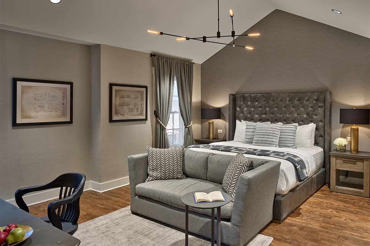 Modern bedroom with a tufted bed, gray sofa, side tables, lamps, wall art, and a contemporary chandelier.