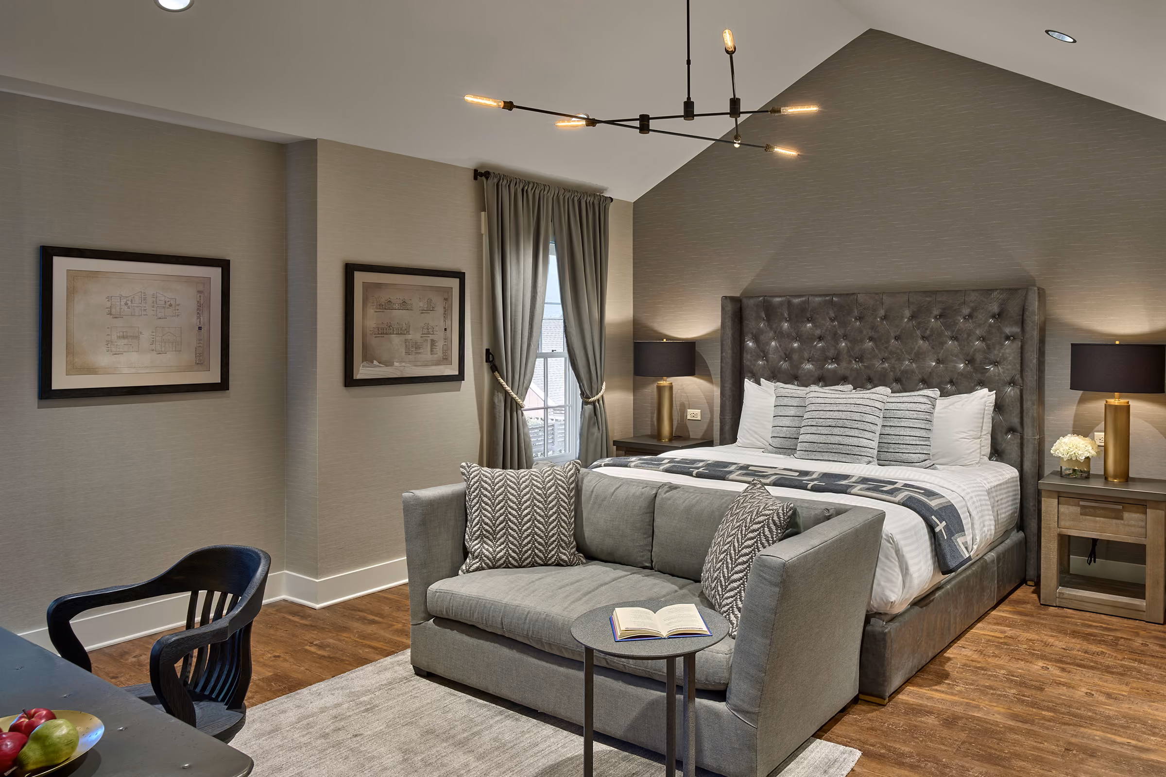Modern bedroom with a tufted bed, gray sofa, side tables, lamps, wall art, and a contemporary chandelier.