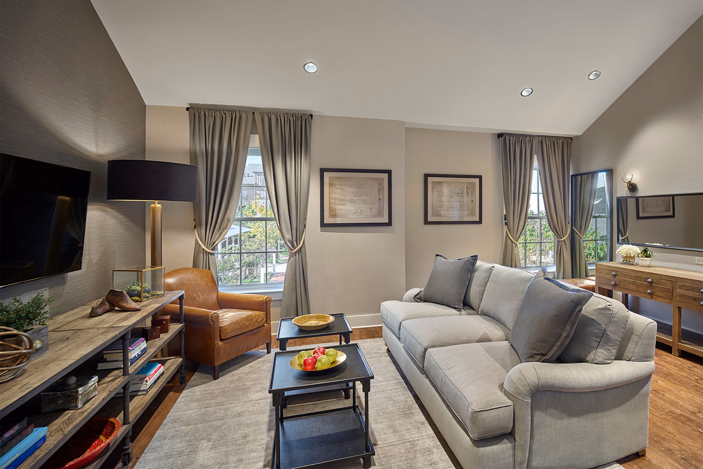 A living room with a gray sofa, leather armchair, coffee tables, TV, bookshelf, and windows with gray curtains.
