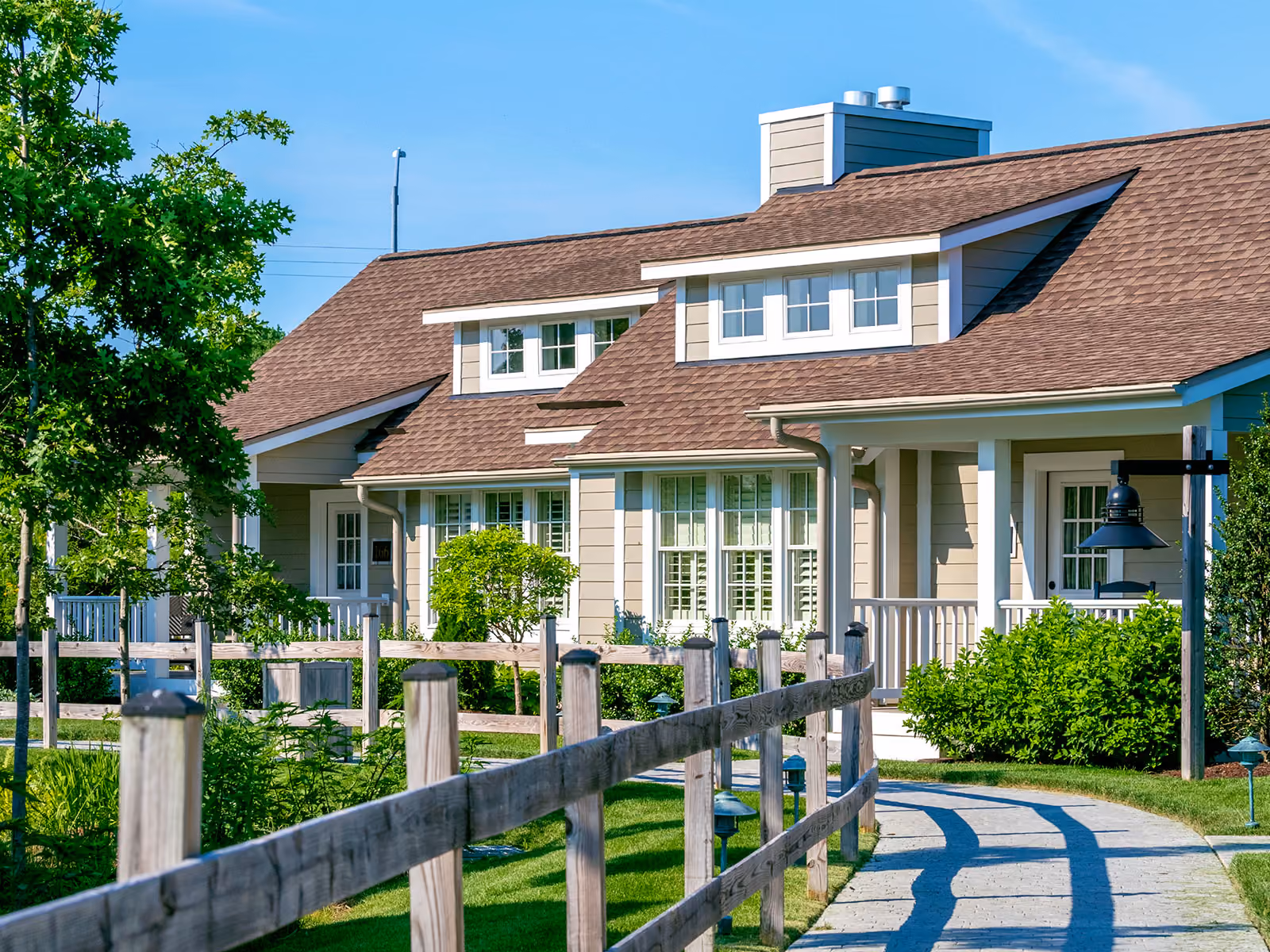 A modern suburban house with a brown roof, large windows, front porch, white fence, and green landscaping.