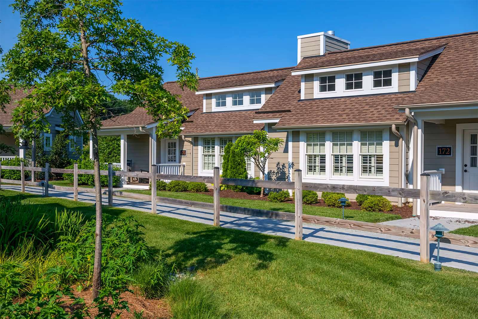 Single-story suburban house with brown roof, beige siding, white trim, and landscaped front yard on a sunny day.