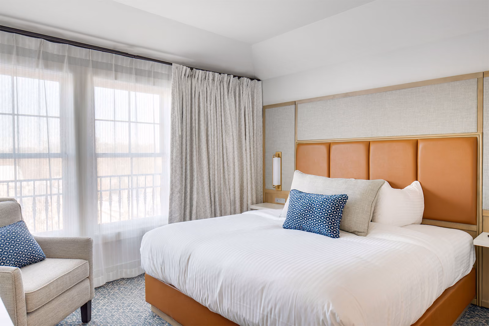 A neatly made hotel bed with white linens and blue accent pillows, next to a window with sheer curtains and an armchair.