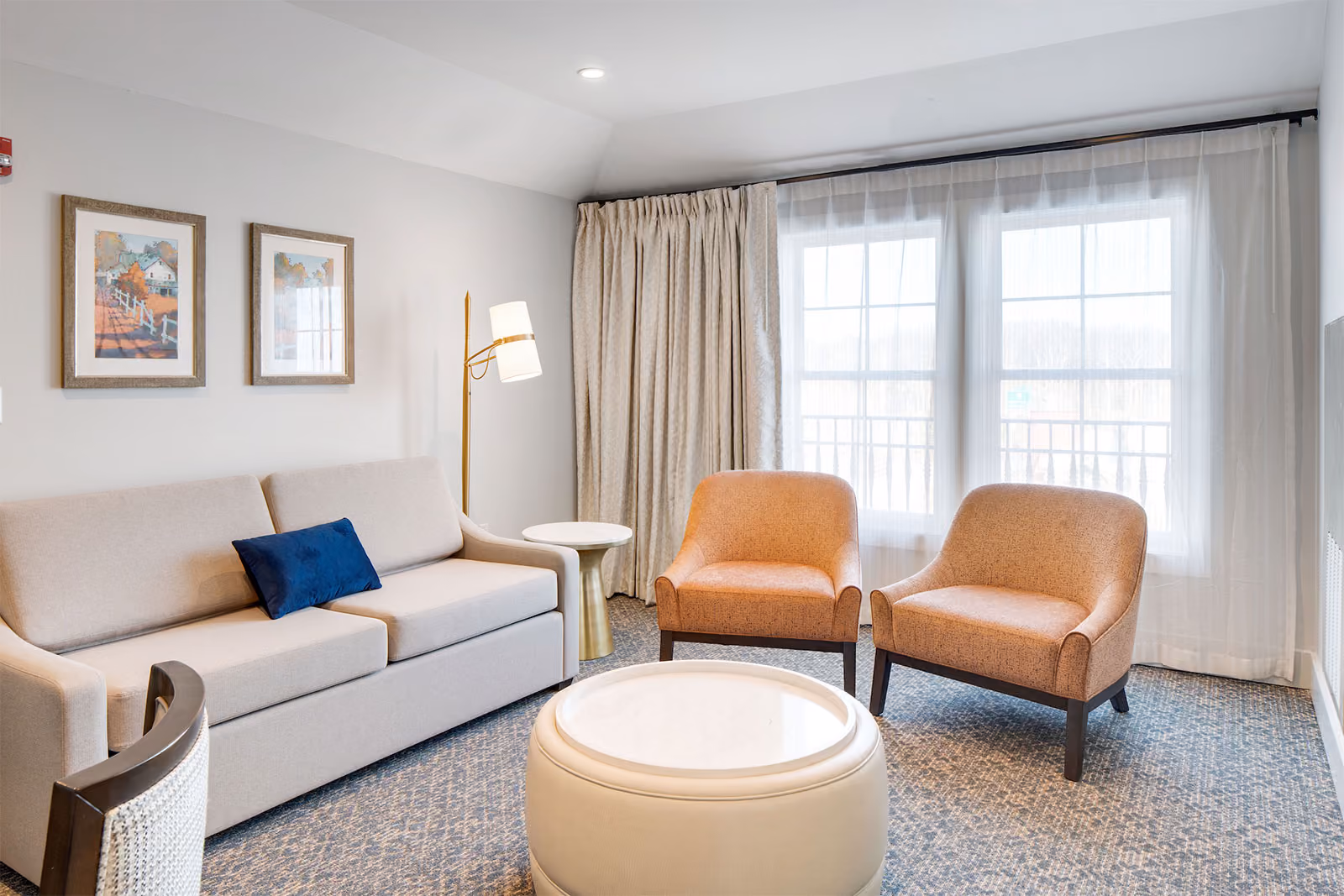 A modern living room with a sofa, two armchairs, round ottoman, framed art, and large windows with sheer curtains.