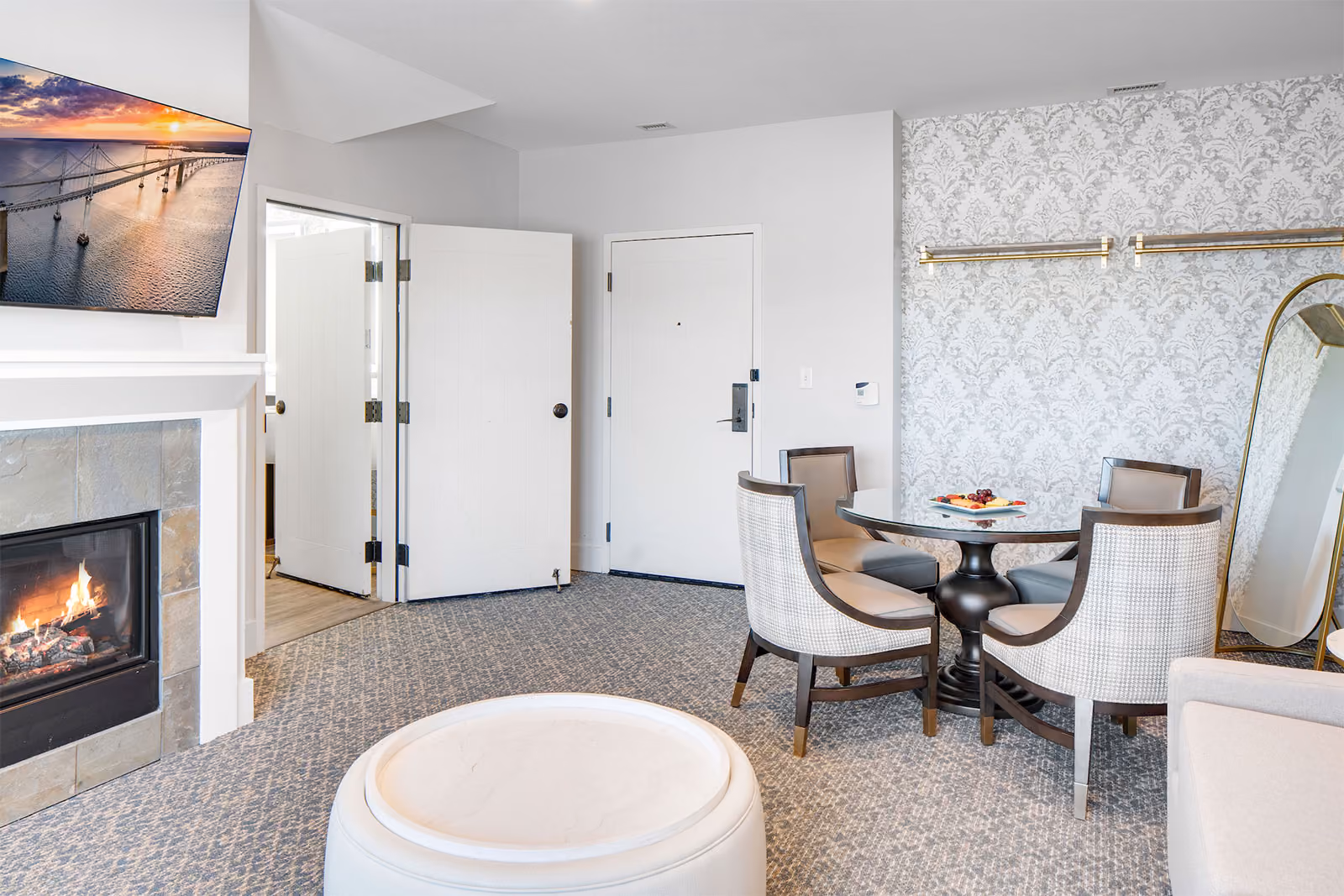 Modern hotel room with a round table, four chairs, fireplace, wall-mounted TV, and decorative wallpaper.