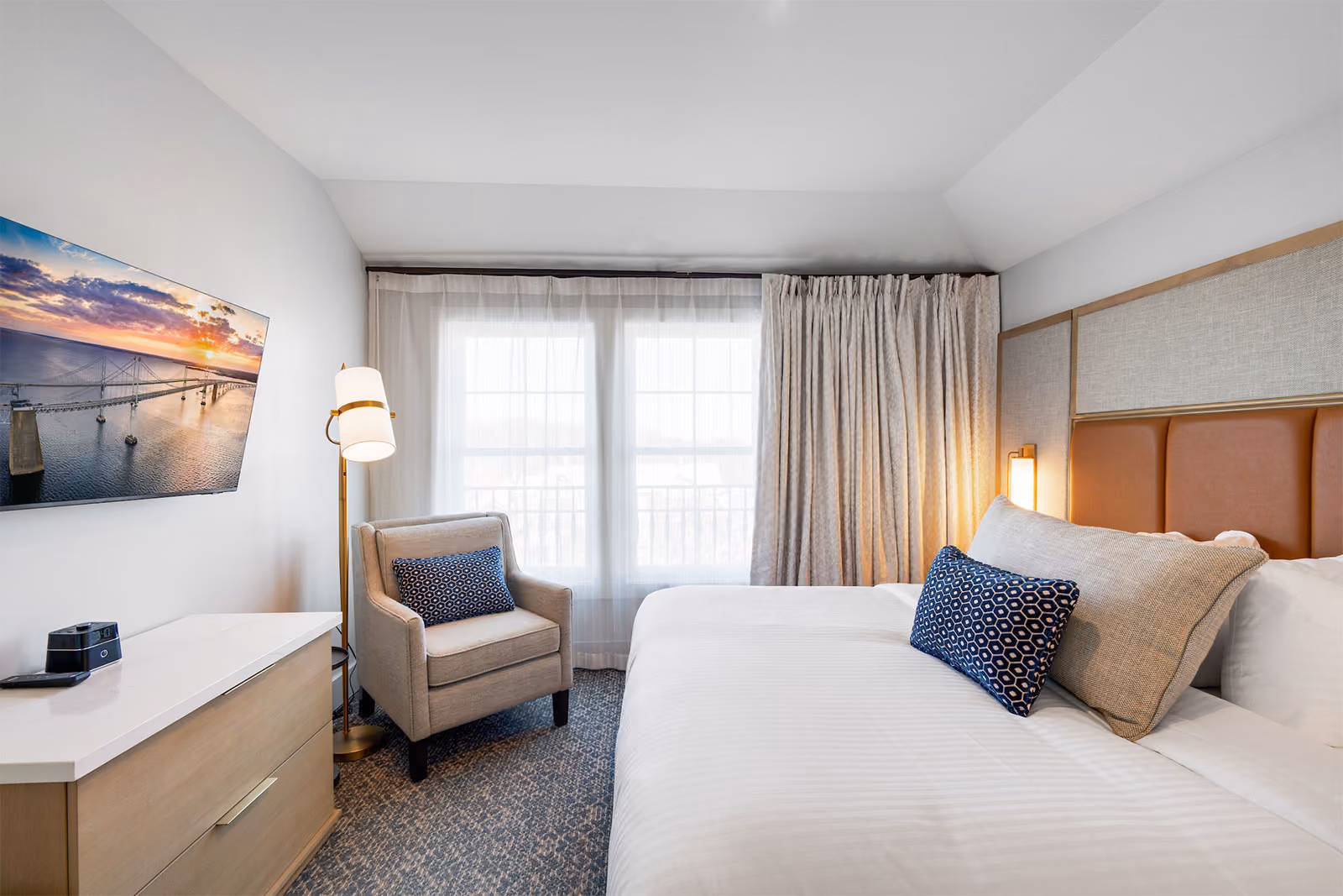 Modern hotel room with a neatly made bed, armchair, TV on the wall, and large window with curtains.