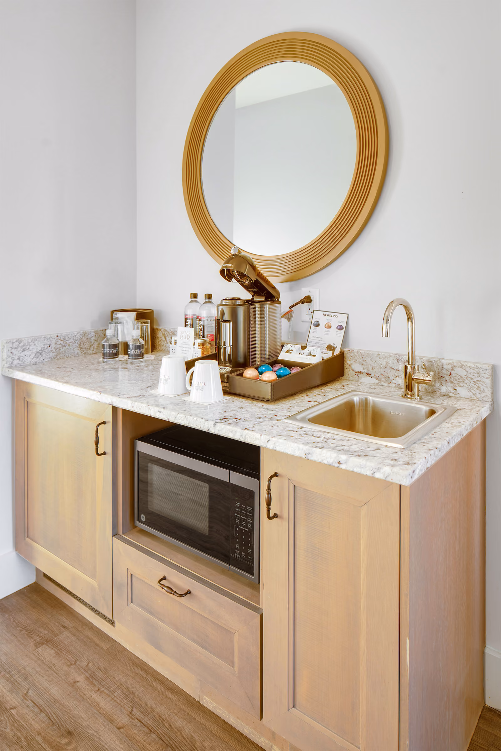 Small kitchenette with a round mirror, coffee station, microwave, sink, and light wood cabinets on a wood floor.