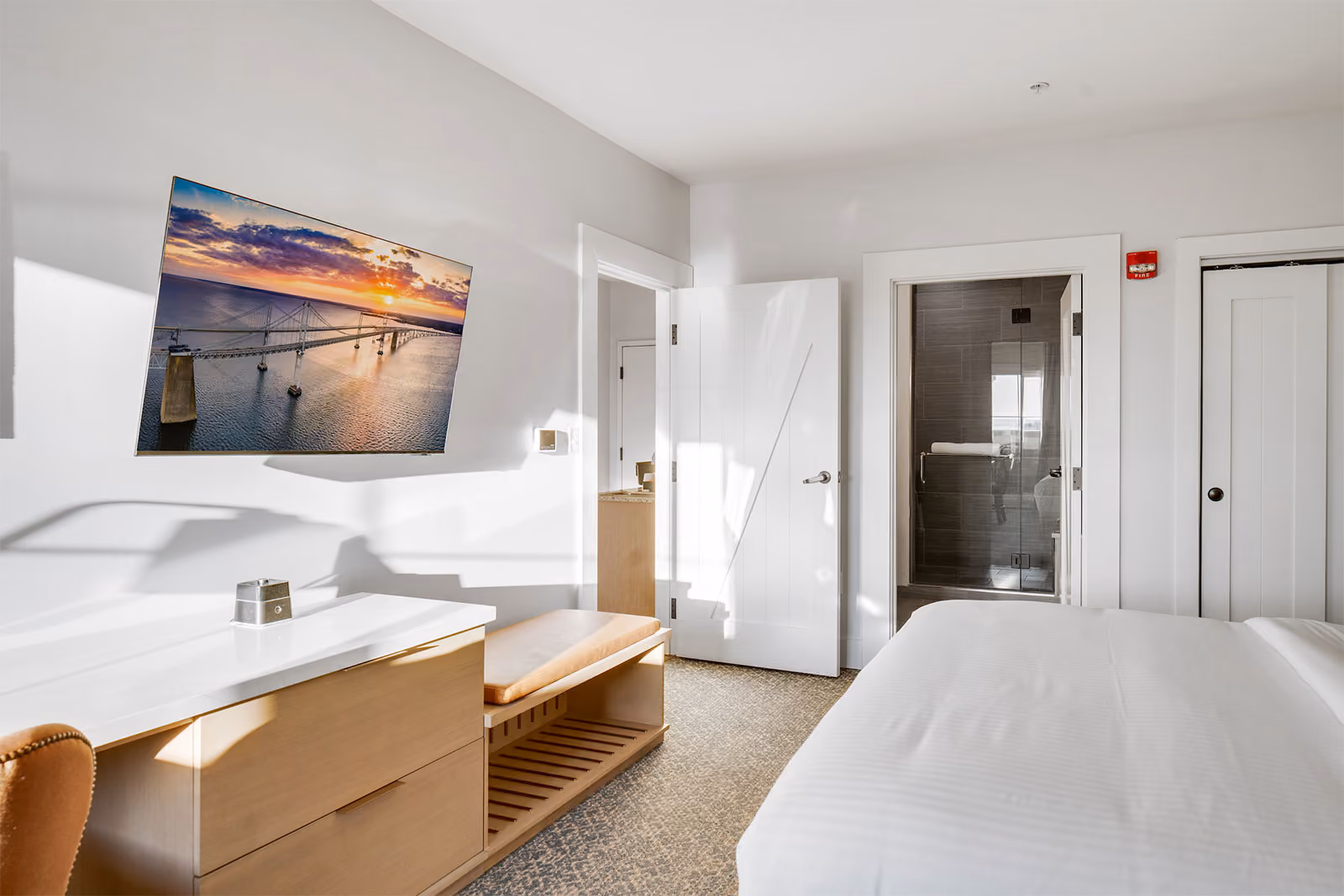 Modern hotel room with a bed, desk, TV showing a bridge, open door to bathroom, and neutral decor.
