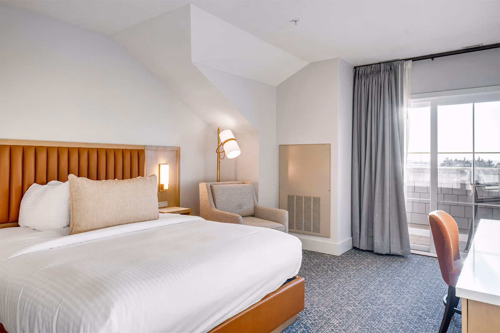 Hotel room with a neatly made bed, armchair, desk, and a sliding glass door leading to a balcony.