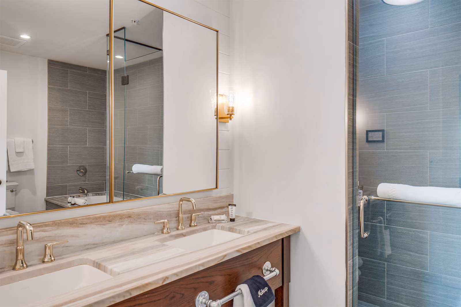 Modern bathroom with double sink, gold fixtures, large mirror, and a glass-enclosed shower with gray tile walls.
