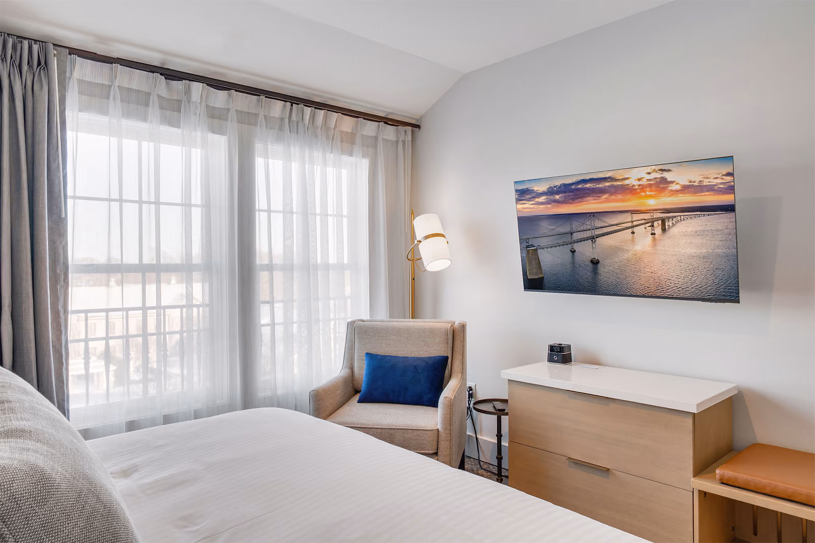 Hotel room with a bed, armchair, dresser, TV displaying a sunset, and large window with sheer curtains.