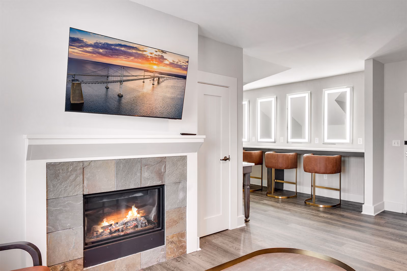 Modern living space with a wall-mounted TV above a fireplace and a bar area with brown chairs in the background.