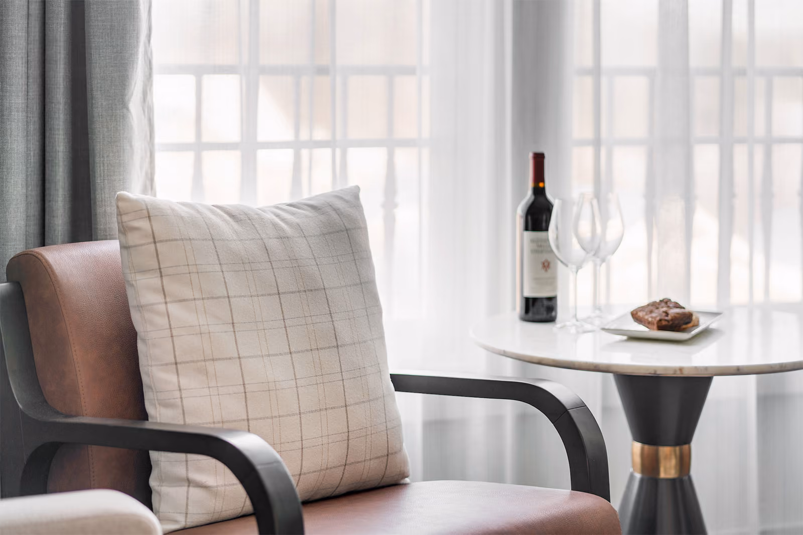 A cushioned chair beside a round table with a wine bottle, two glasses, and a pastry near a window with sheer curtains.