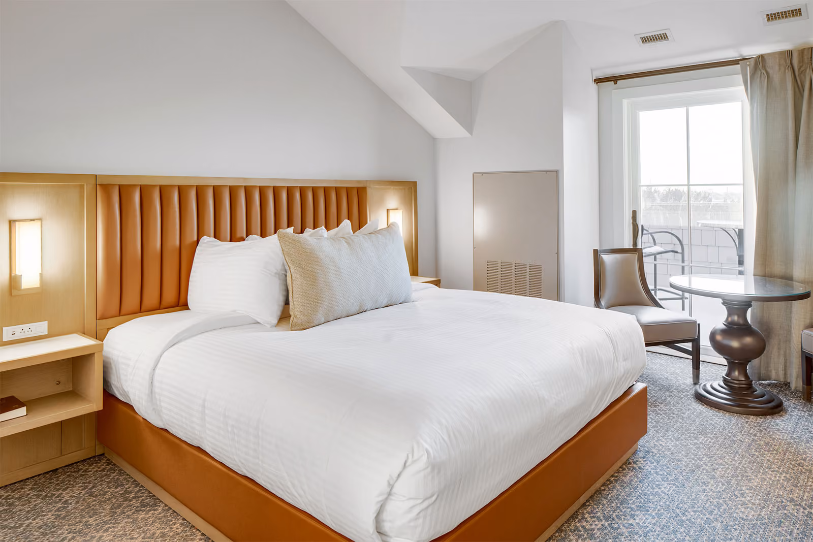 Hotel room with a large bed, white linens, bedside tables, a chair, and a round table near a window.