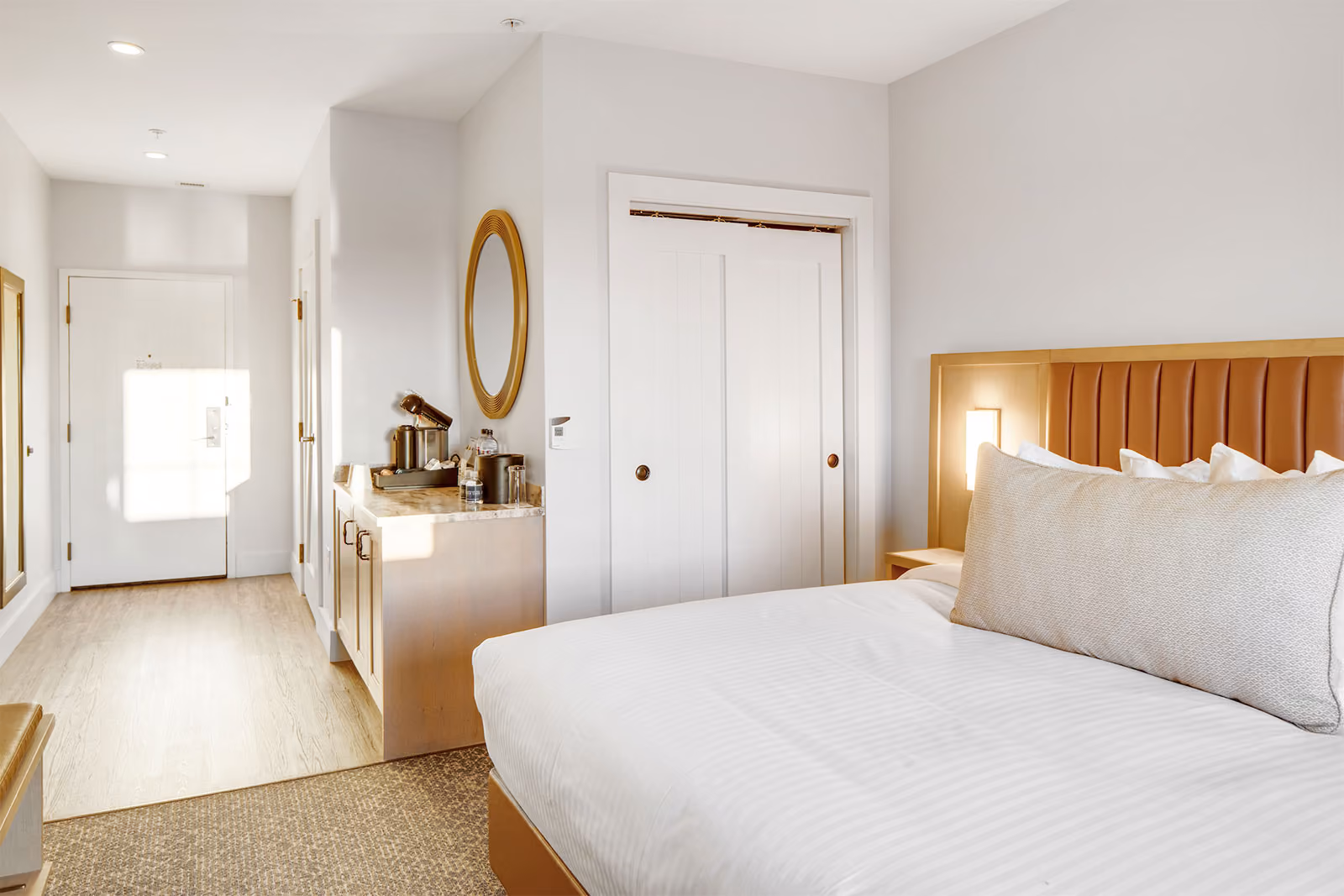 Modern hotel room with a neatly made bed, a wooden headboard, a vanity area, and white walls.