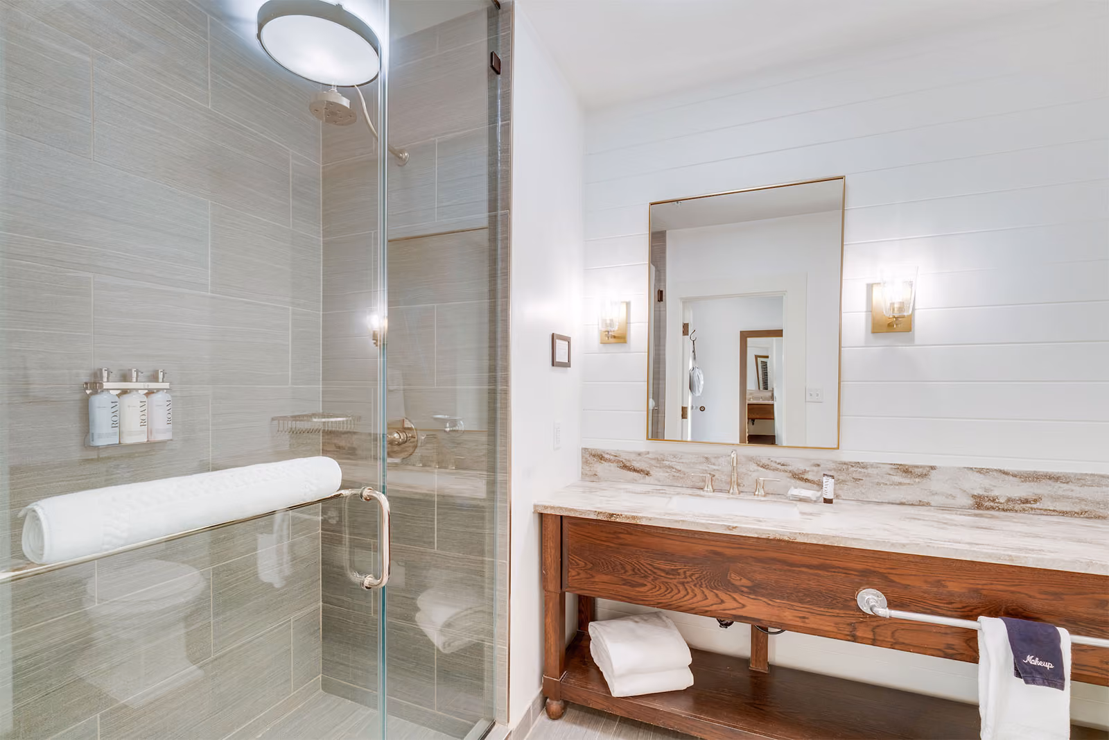 Modern bathroom with a glass shower, wall-mounted toiletries, large mirror, marble countertop, and wooden vanity.