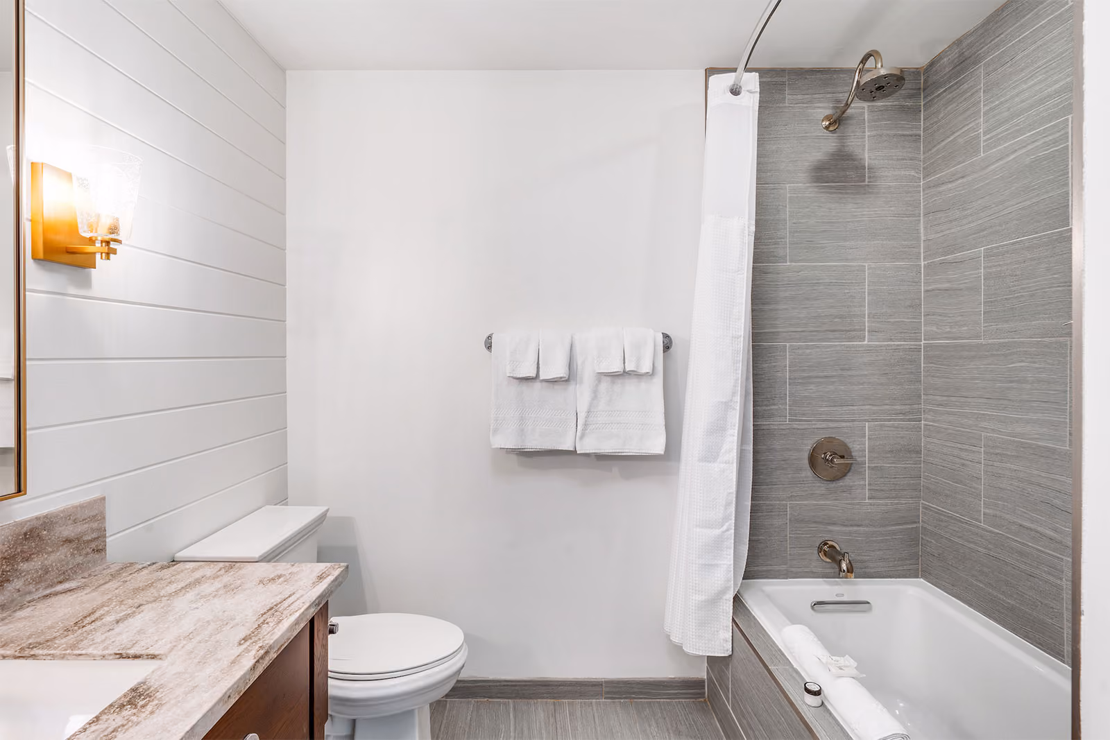 Modern bathroom with gray tile tub-shower, white curtain, toilet, vanity with marble countertop, and white towels.