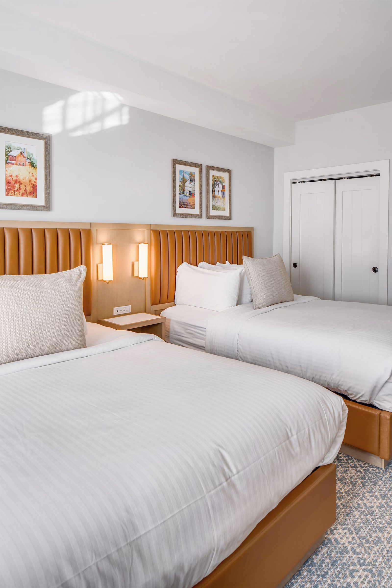 Two neatly made beds with tan headboards in a bright, modern bedroom with framed art and white double closet doors.