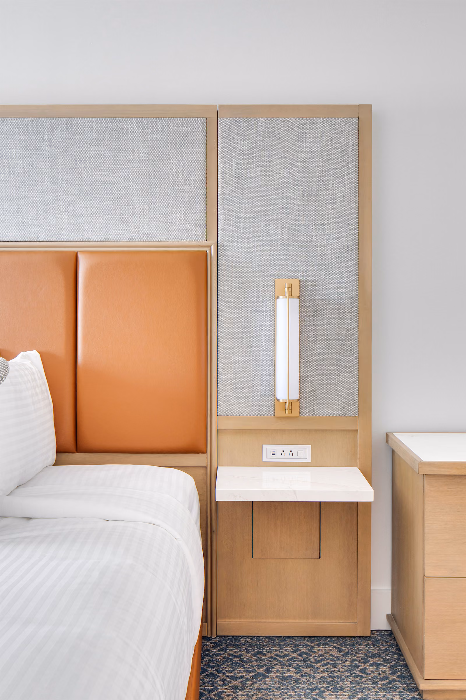 Modern hotel room nightstand with a lamp, power outlets, and part of a bed with striped white bedding.