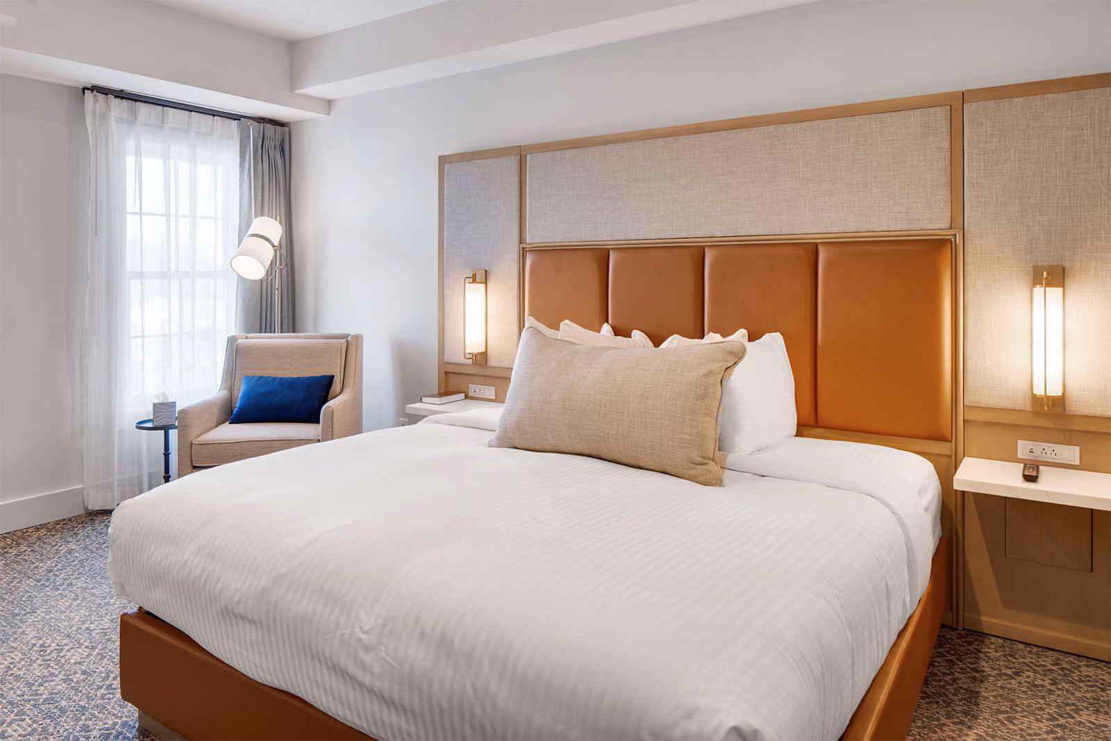 Modern hotel room with a large bed, tan headboard, armchair by the window, and bedside lamps.