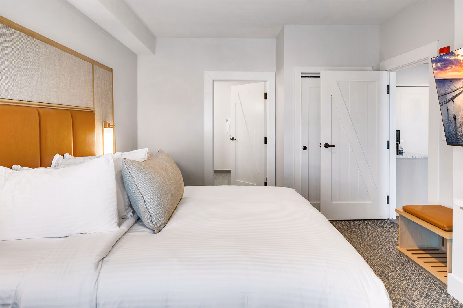Hotel room with neatly made bed, white linens, padded headboard, open doors, and wall-mounted TV.