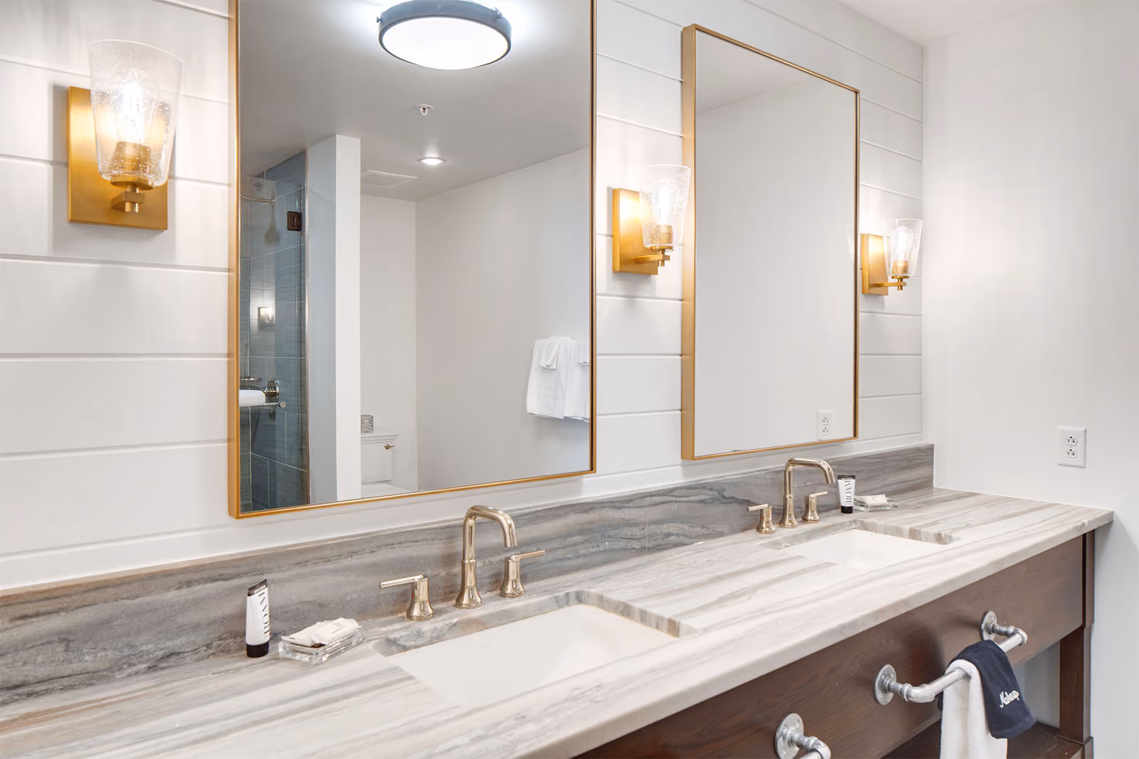 Modern bathroom with double sinks, marble countertop, gold fixtures, two mirrors, and wall-mounted lights.