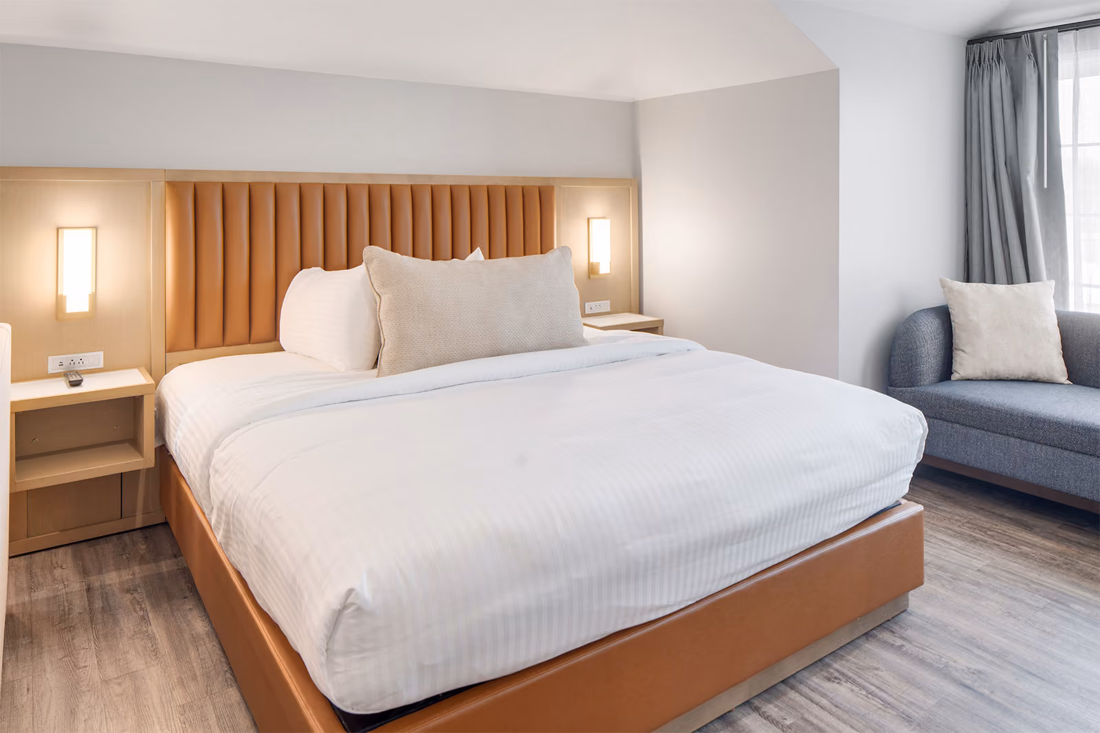 A modern hotel room with a large bed, two bedside tables, wall lights, and a gray sofa by a window.