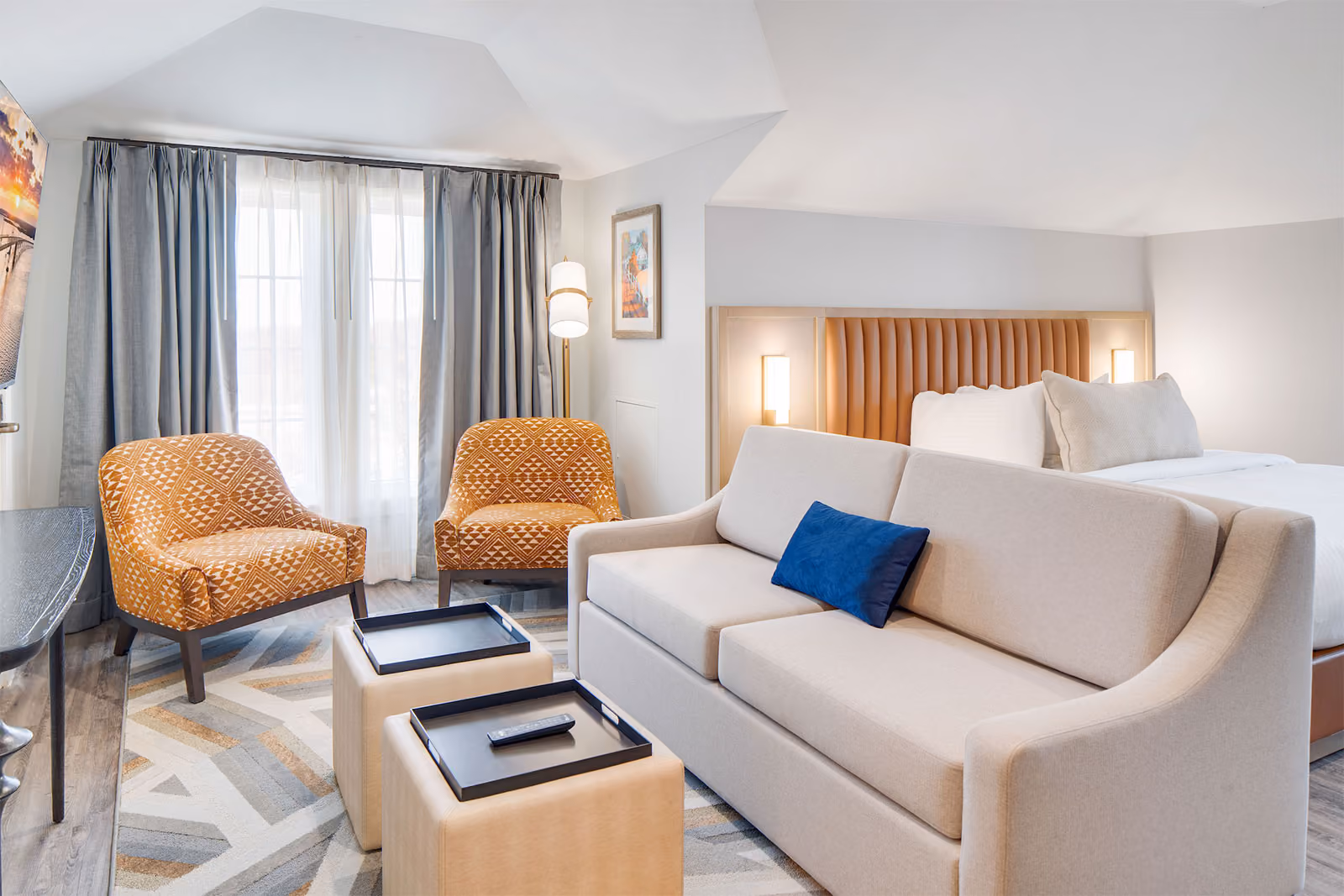 Modern hotel room with a bed, sofa, two patterned chairs, side tables, and large windows with curtains.