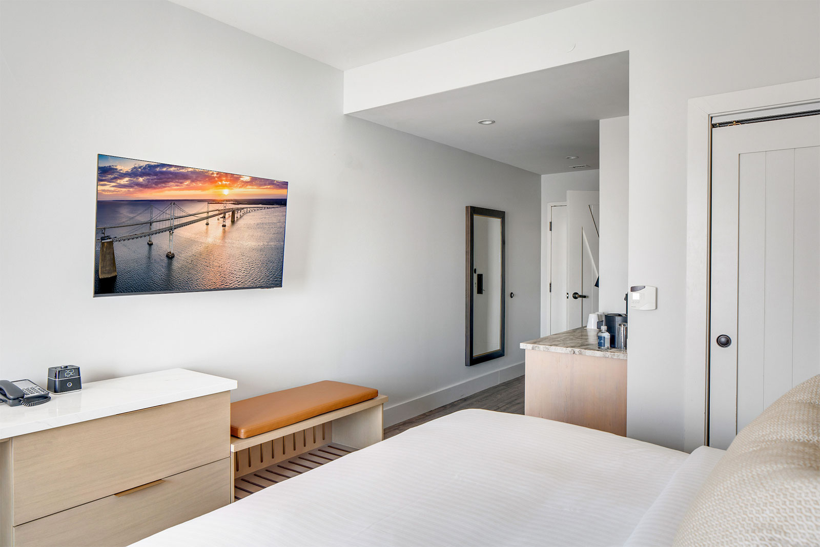 Modern hotel room with white walls, a bed, a wall-mounted TV displaying a sunset, and minimalist furnishings.