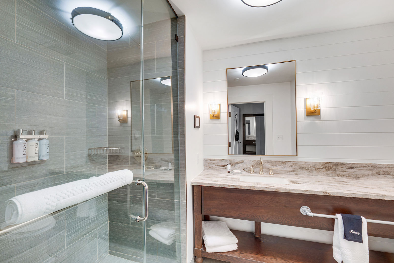 Modern bathroom with glass shower, light wood vanity, large mirror, wall sconces, and neatly folded towels.