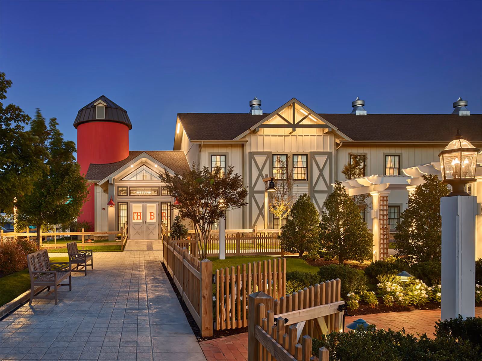 A farmhouse-style building with a red silo, lit at dusk, surrounded by gardens, benches, and a wooden fence.