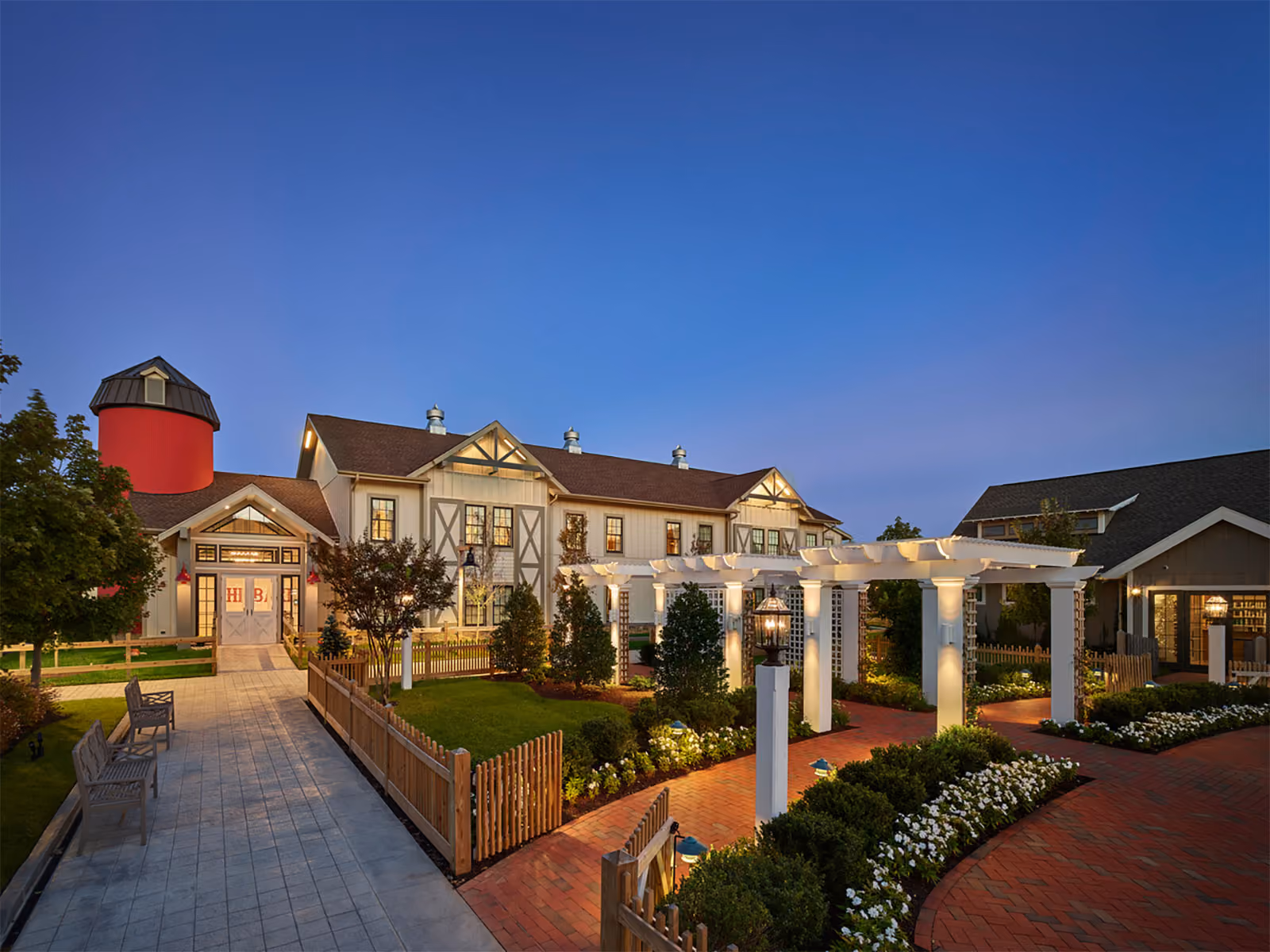 Evening view of a large barn-style building with a red silo, landscaped gardens, and a white pergola walkway.