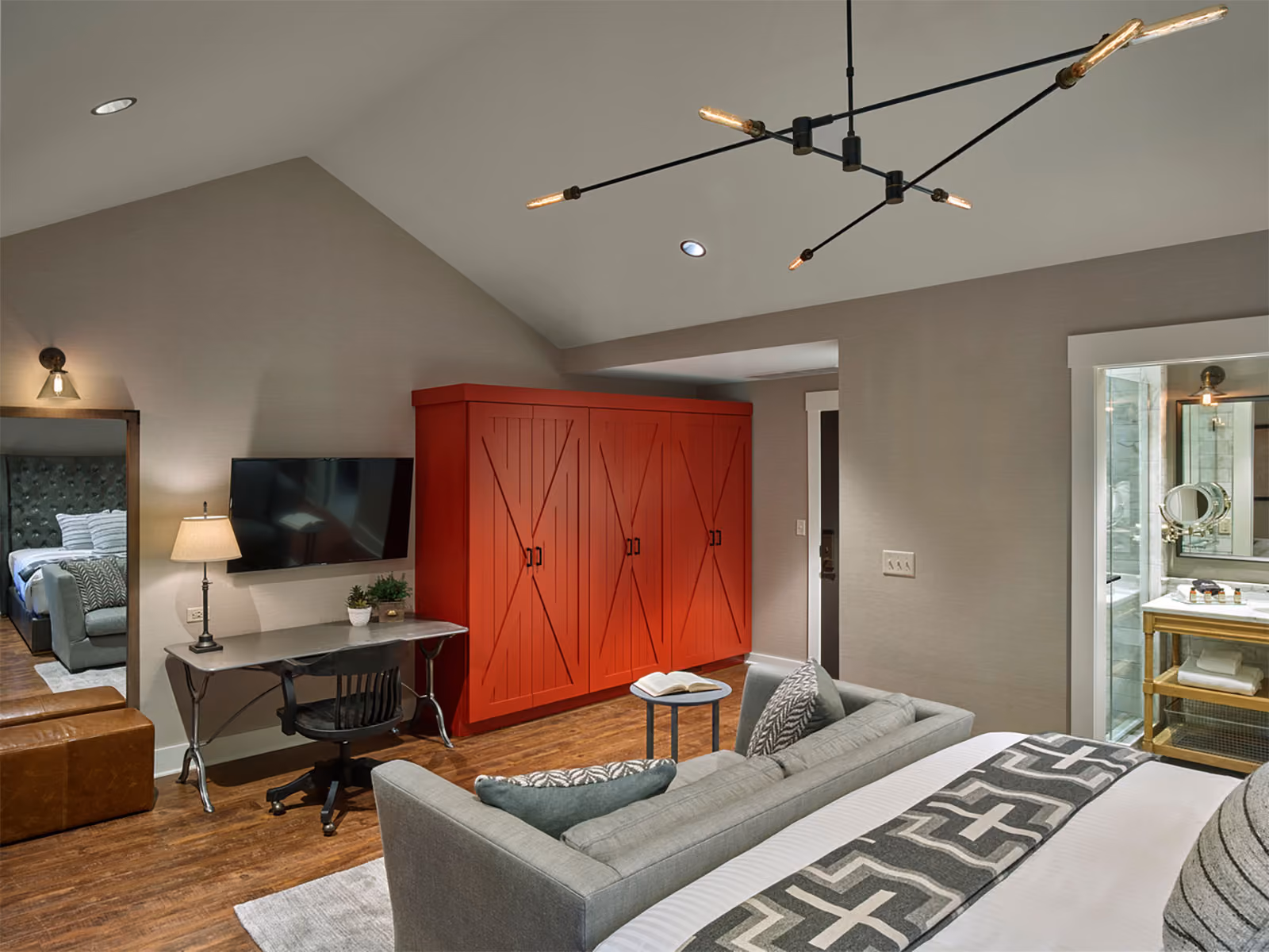 Modern bedroom with a red wardrobe, desk, wall-mounted TV, and open view into bathroom and adjoining room.
