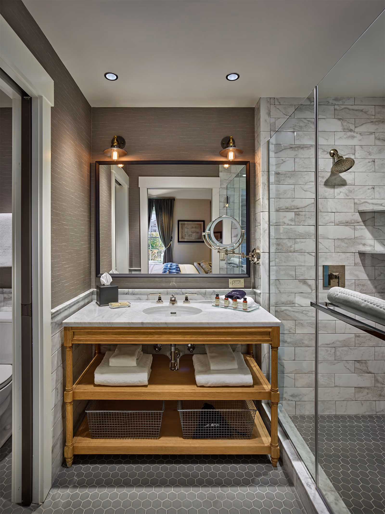 Modern bathroom with a marble vanity, large mirror, glass shower, and open shelves with towels and baskets underneath.