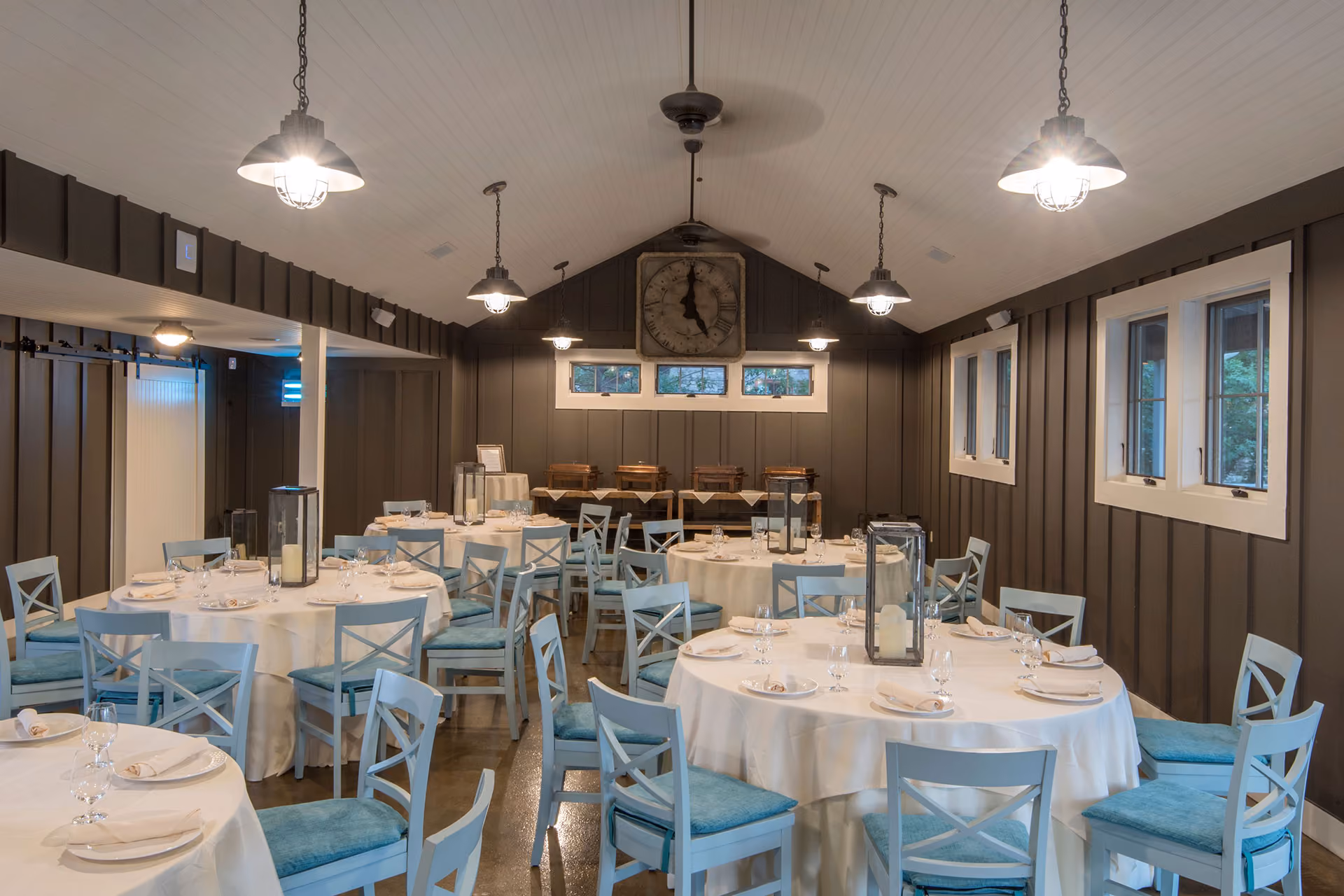 A dining room with round tables, blue chairs, place settings, lantern centerpieces, and overhead lights.