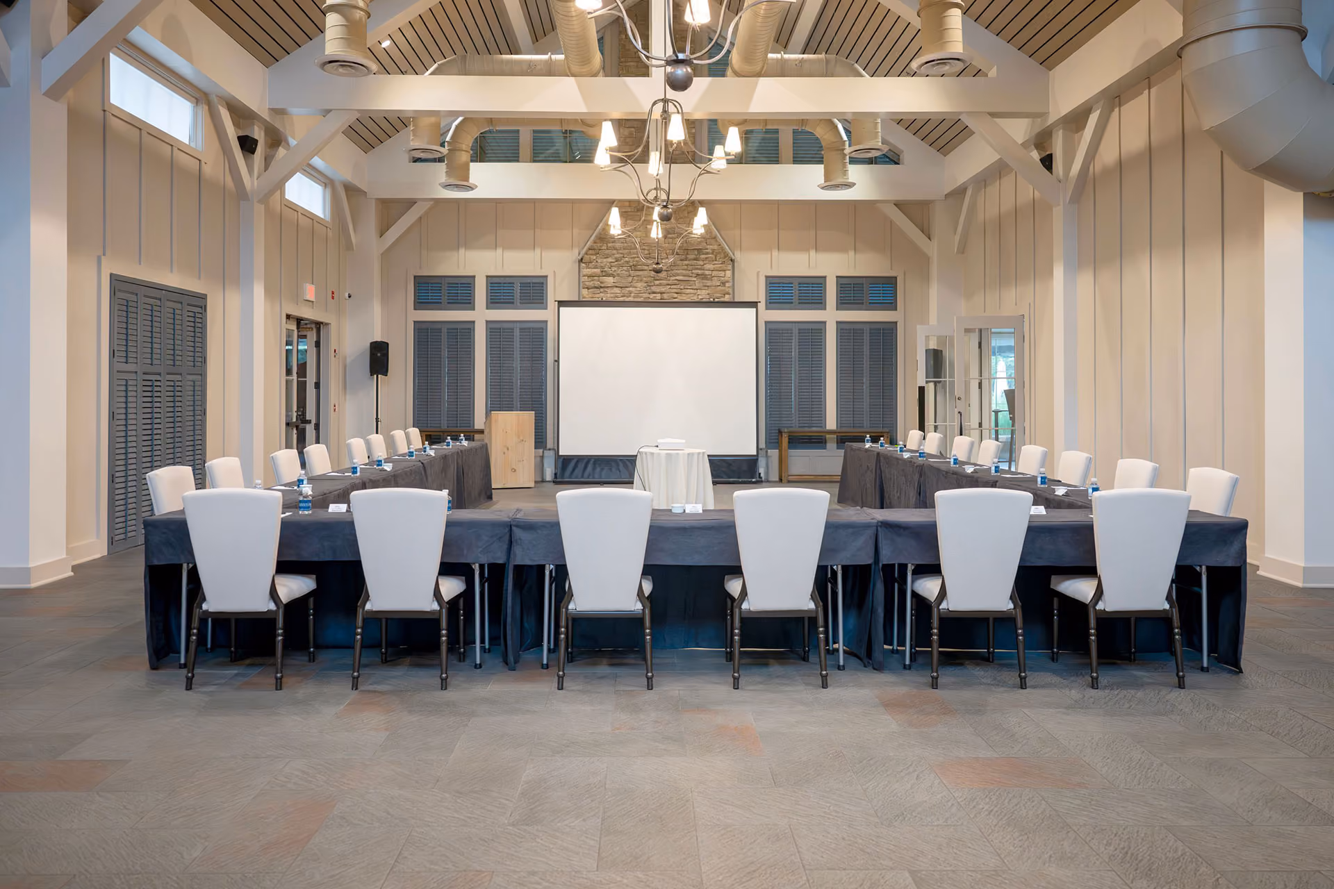 Conference room with U-shaped table setup, white chairs, a projector screen, and modern ceiling fixtures.