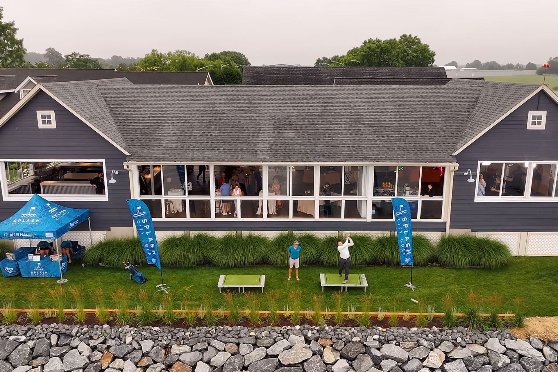 Aerial view of two people playing golf on grass outside a building with large windows and blue event banners.